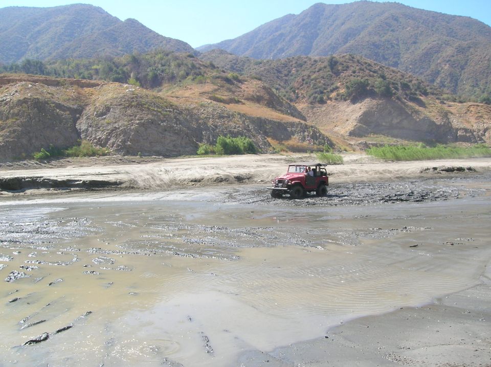 Red off-road vehicle driving through a muddy, dry riverbed with hills in the background