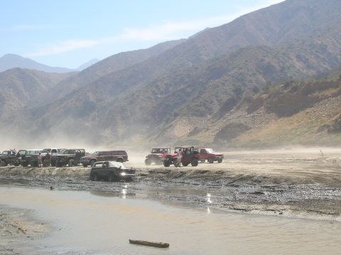 1985 Ford Bronco - A group of off-road vehicles navigates a challenging dusty and muddy environment, with a mountainous backdrop.