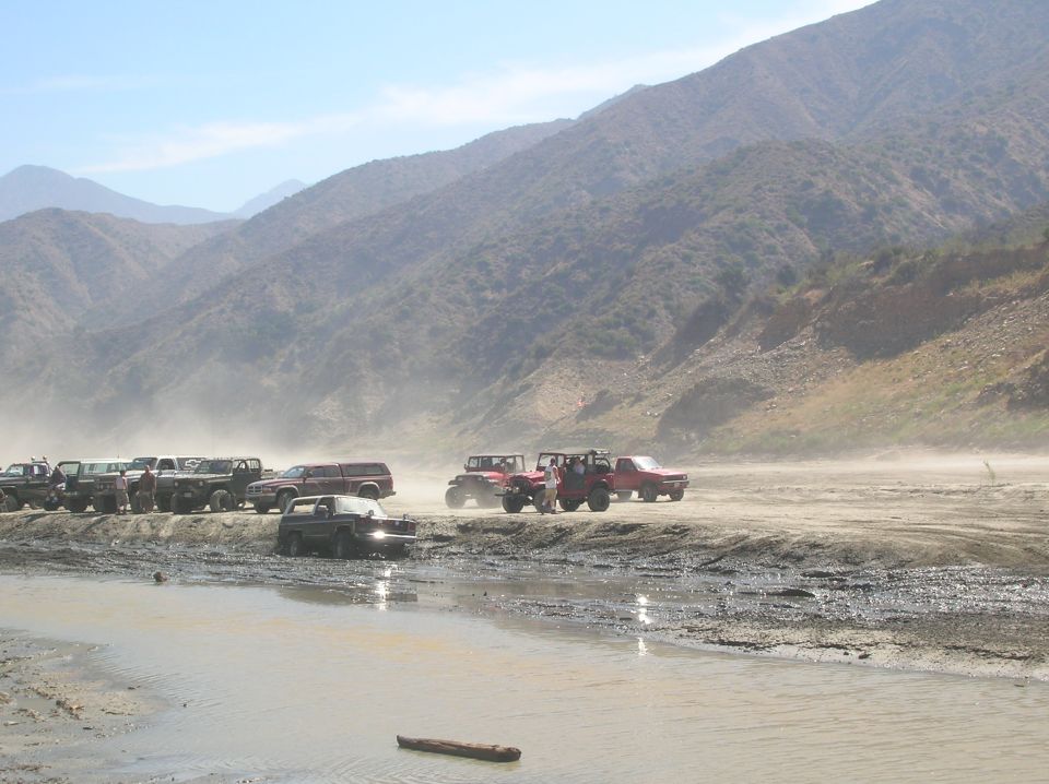 Multiple off-road vehicles gathered in a dusty, muddy terrain with mountains in the background