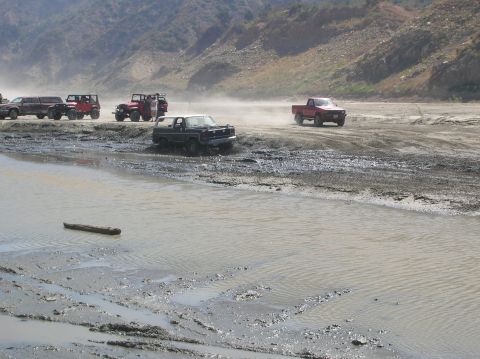 1985 Ford Bronco - Multiple off-road vehicles traverse a challenging muddy and watery landscape under a bright sky.