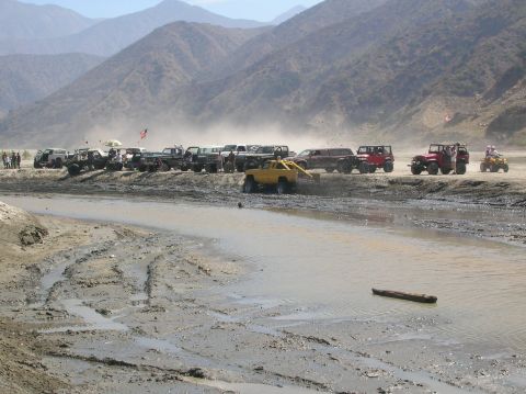 1985 Ford Bronco - Multiple off-road vehicles are gathered on a dirt track next to a muddy water area with mountains in the distance.