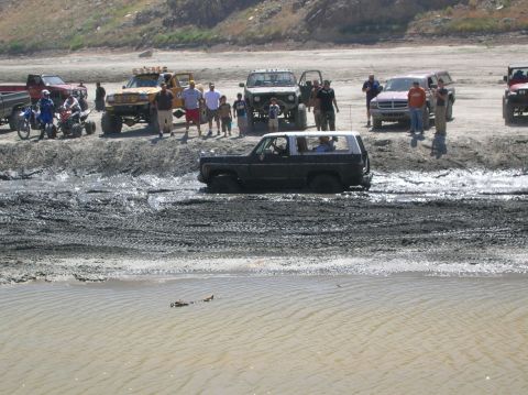 1985 Ford Bronco - A group of off-road vehicles are gathered around as a black 1985 Ford Bronco struggles through a muddy track.