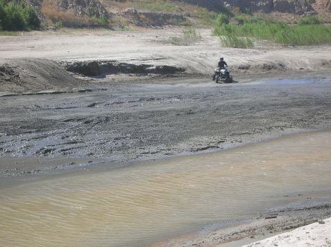 1985 Ford Bronco - A person rides an ATV through a muddy, dry riverbed with sandy banks and vegetation.
