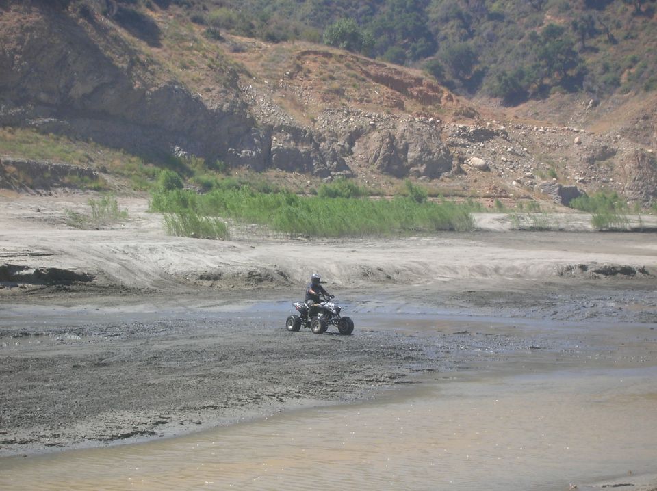 Quad bike with rider on muddy terrain near a body of water, with hills in the background