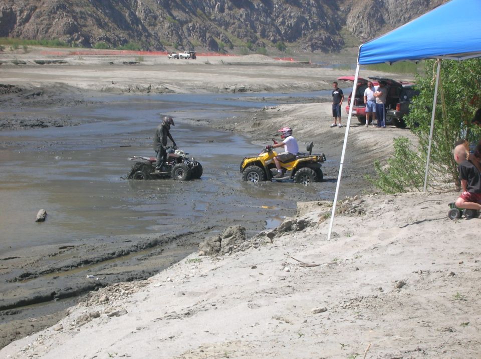Two ATVs stuck in mud, with people watching from a sandy bank under a blue canopy