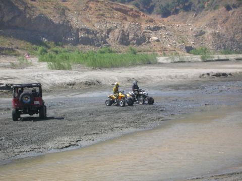 1985 Ford Bronco - A red Jeep Wrangler is parked on a muddy surface next to a body of water, with two ATVs in the distance.