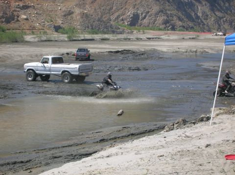 1985 Ford Bronco - A white pickup truck navigates a muddy terrain, while an ATV creates a splash nearby. Other vehicles and a blue tent are visible in the bac…