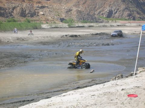 1985 Ford Bronco - Multiple off-road vehicles navigate a muddy, dry riverbed under a bright sun.