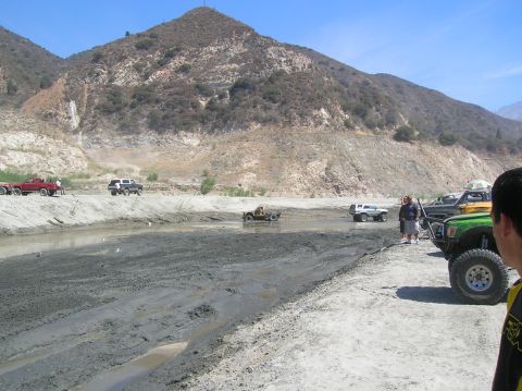 1985 Ford Bronco - Off-road vehicles navigate a challenging muddy terrain under a clear blue sky with mountainous backdrop.