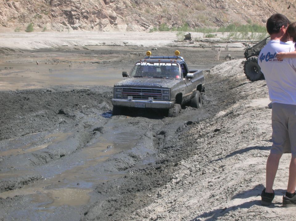 Black pickup truck with yellow lights on roof rack stuck in mud, another off-road vehicle visible in background