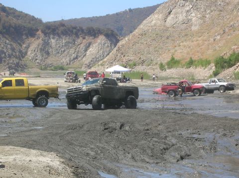 1985 Ford Bronco - Several pickup trucks are stuck in the mud at an off-road park, with spectators watching from the sidelines.