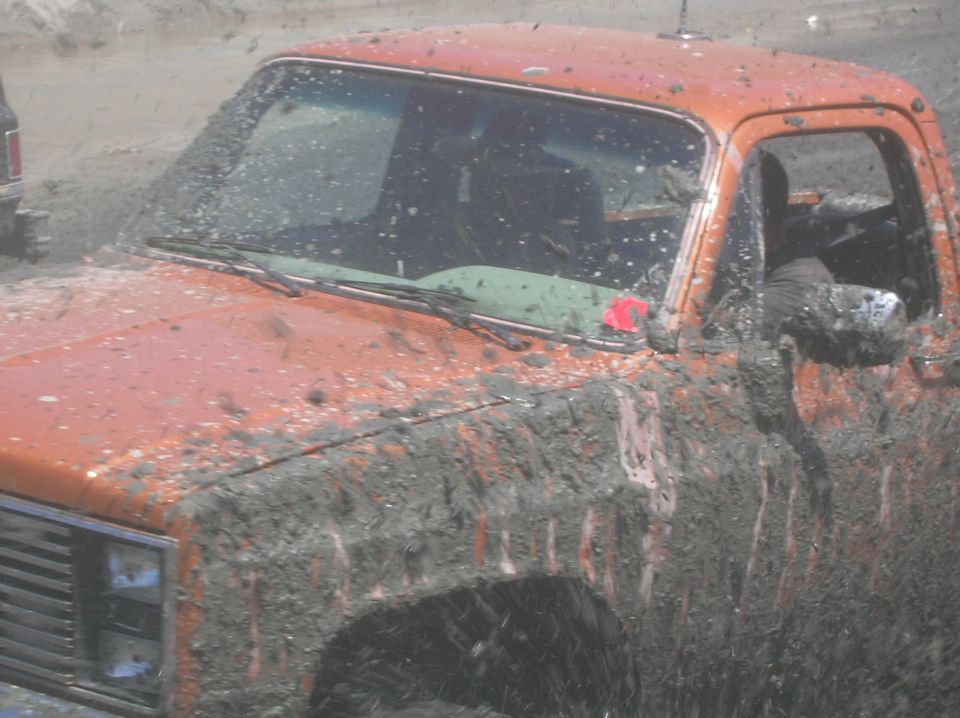 Mud-covered orange pickup truck, front fender and door visible. Another vehicle is partially visible to the left
