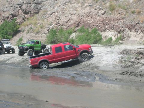 1985 Ford Bronco - Several off-road vehicles navigate a muddy terrain with rocky hills in the background.