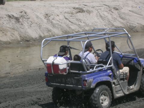 1985 Ford Bronco - Three people ride in a blue off-road vehicle with a roll cage as it travels on a muddy path beside a body of water.