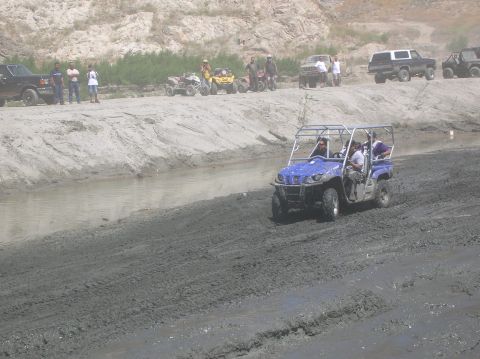 1985 Ford Bronco - A blue UTV navigates a muddy off-road course, with other vehicles and people visible in the background.