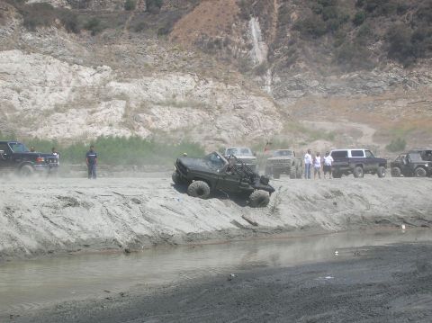 1985 Ford Bronco - Off-road vehicles navigate a dusty, sandy area with a muddy stream and rocky hills.