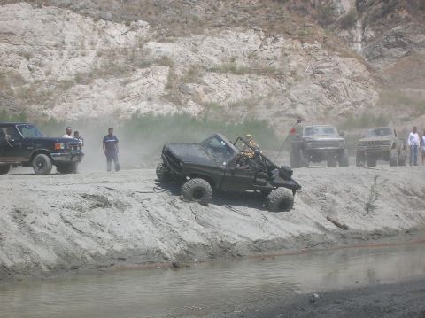 1985 Ford Bronco - Off-road vehicles navigate a dusty trail near a riverbed with spectators watching.