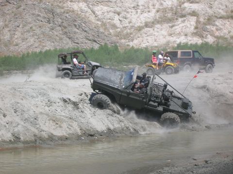 1985 Ford Bronco - Off-road vehicles traverse a challenging muddy track, kicking up dust and water.