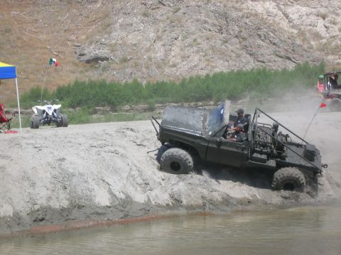 1985 Ford Bronco - A modified off-road vehicle navigates a muddy bank next to a body of water, with an ATV visible in the background.
