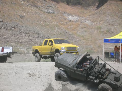 1985 Ford Bronco - Multiple off-road vehicles navigate a dirt path near a rocky hillside, with a yellow truck and a modified vehicle in the foreground.