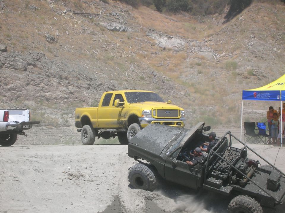 Yellow Ford F-Series truck and a muddy off-road vehicle with people inside, on a dirt path with a rocky hill in the background