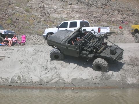 1985 Ford Bronco - A heavily modified off-road vehicle with its engine exposed sits on a sandy bank near the water, with other vehicles and people in the back…