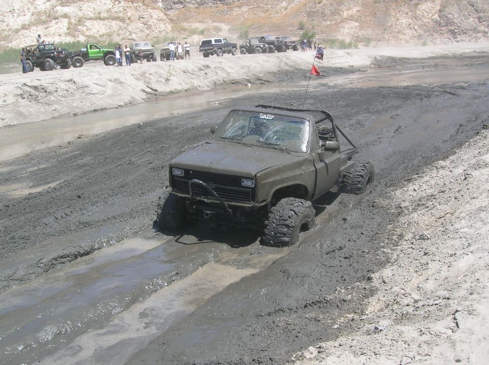 modified pickup truck with large tires stuck in mud, other vehicles and people in background