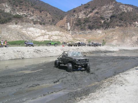 1985 Ford Bronco - Several off-road vehicles navigate a muddy terrain with mountains in the background.
