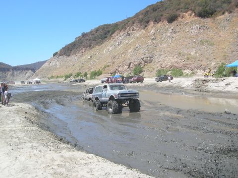 1985 Ford Bronco - Off-road vehicles navigate a muddy terrain under a clear blue sky, with spectators gathered on the bank.