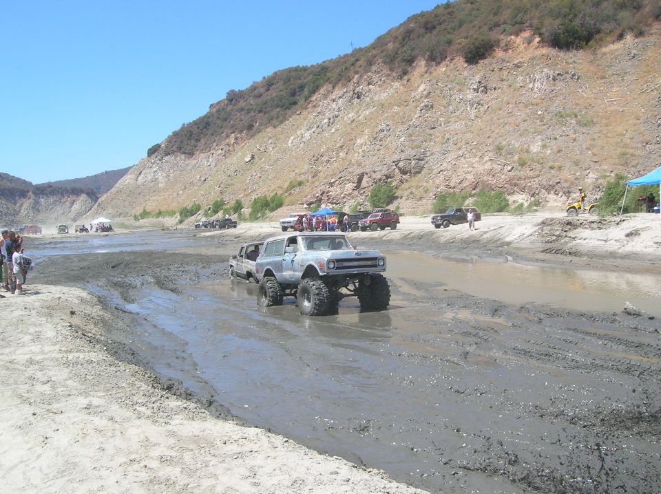 Lifted off-road SUV with large tires driving through mud, other vehicles and spectators in background