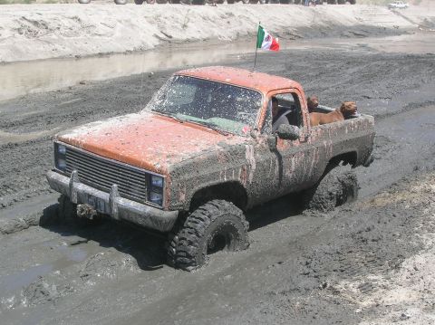 1985 Ford Bronco - An orange pickup truck is stuck in thick mud, with a Mexican flag flying from its roof.
