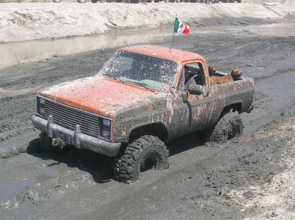 Orange pickup truck with large tires stuck in mud, front grille visible, Mexican flag on roof