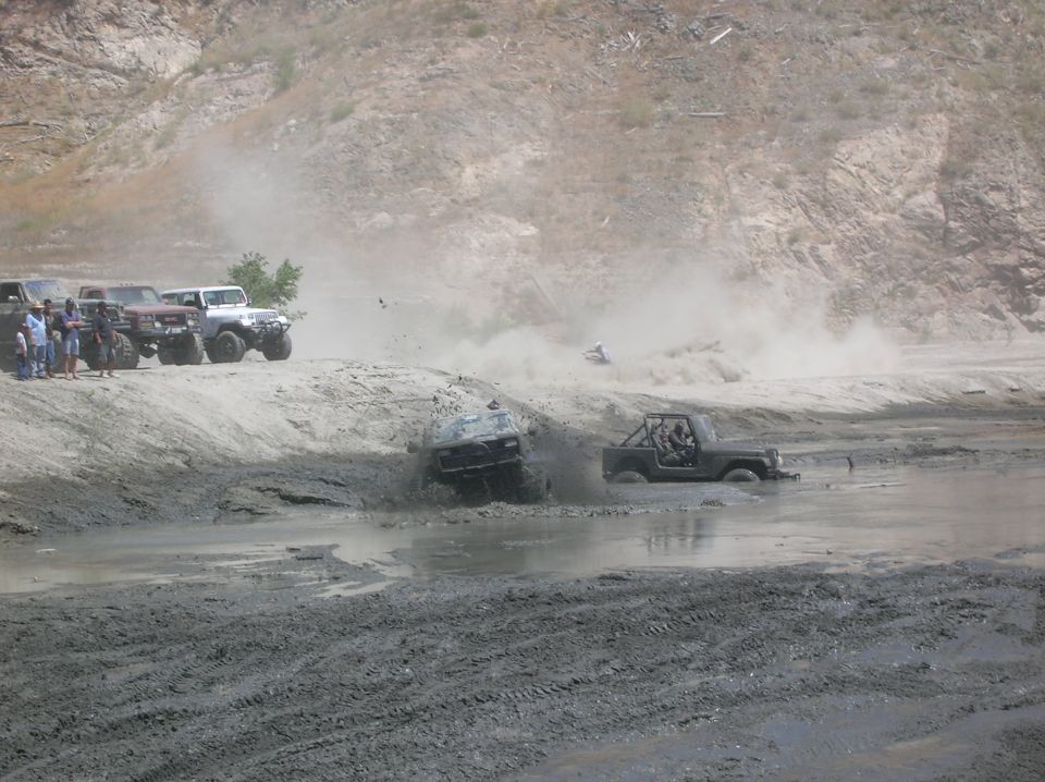 Several off-road vehicles are navigating a mud pit, with one vehicle partially submerged and kicking up mud