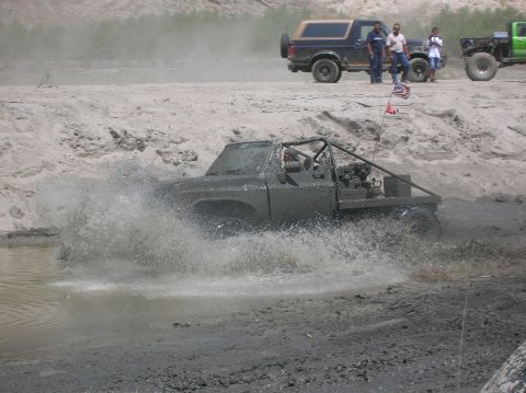 1985 Ford Bronco - A heavily modified pickup truck navigates a muddy water obstacle, kicking up a large spray.