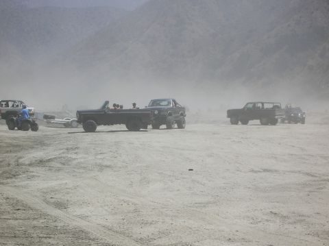 1985 Ford Bronco - Several vehicles are seen in a dusty, outdoor scene, including a black convertible SUV with passengers.