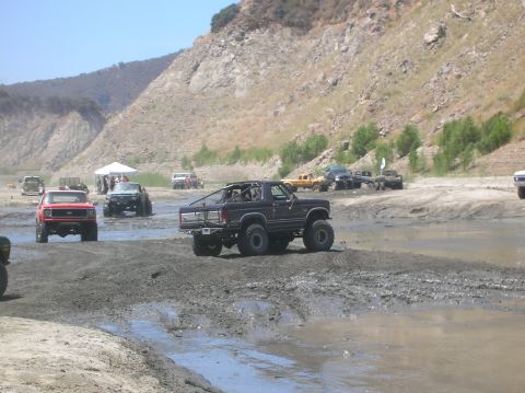 1985 Ford Bronco - Several off-road vehicles navigate a muddy terrain under a bright sky with a rocky hillside.