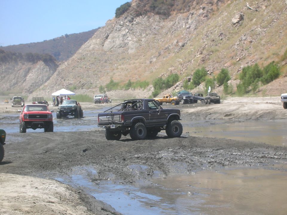 Dark-colored off-road SUV with roll cage driving through mud, other trucks in background