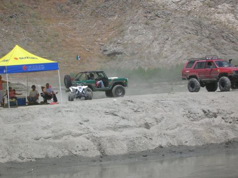 1985 Ford Bronco - A modified green SUV, possibly from the early 1980s, kicks up dust while driving on a sandy terrain next to a red SUV.