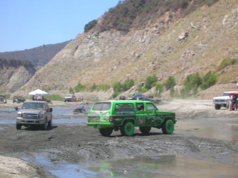 1985 Ford Bronco - Off-road vehicles navigate a muddy terrain under a clear sky with a rocky hillside.