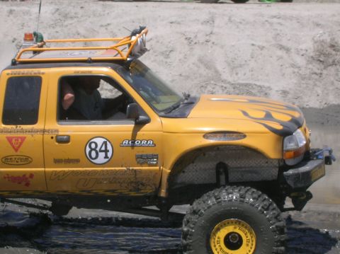 1985 Ford Bronco - A yellow off-road truck navigates through muddy terrain with its driver visible.