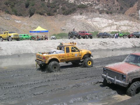 1985 Ford Bronco - Off-road vehicles navigate a muddy track, with a yellow pickup truck prominently featured in the foreground.