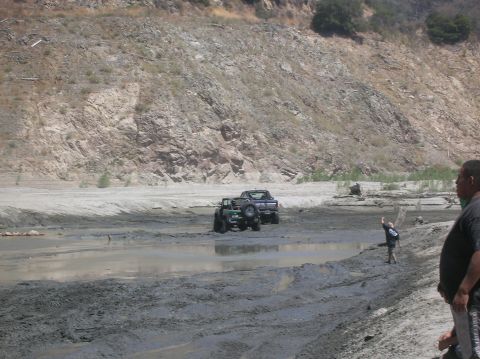 1985 Ford Bronco - Off-road vehicles traverse a muddy terrain under a bright sky, with spectators observing from the bank.