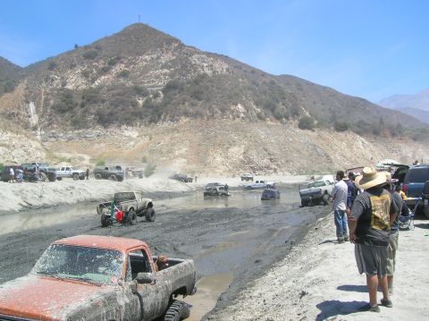 1985 Ford Bronco - Off-road enthusiasts navigate a challenging muddy course, with spectators observing the action from the embankment.