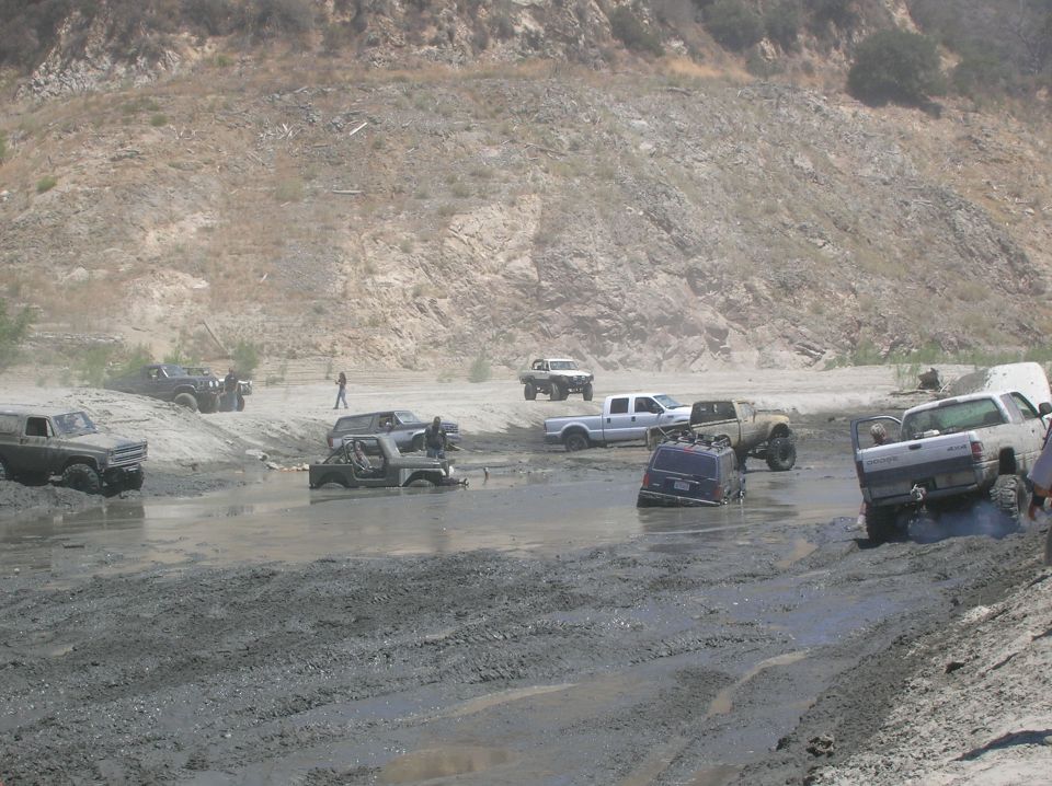 Several off-road vehicles, including trucks and Jeeps, are stuck in deep mud in a dry riverbed with a rocky hillside in the background