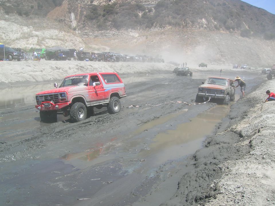 1985 Ford Bronco stuck in mud, another vehicle being pulled out with a rope. Several other vehicles and tents in background