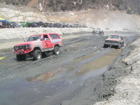 1985 Ford Bronco - Off-road vehicles navigate a muddy track, with one truck being towed.