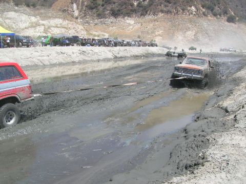 1985 Ford Bronco - Off-road vehicles navigate a muddy track with spectators watching from the embankment.