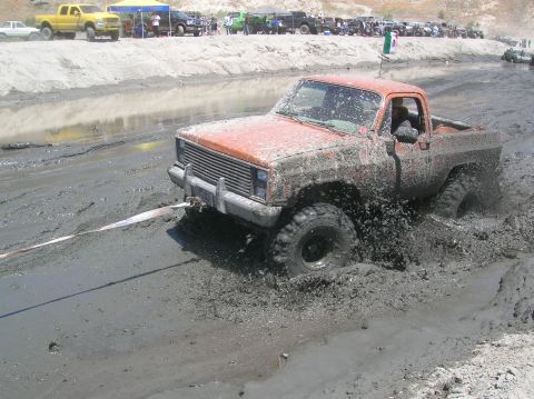 1985 Ford Bronco - An orange pickup truck navigates a muddy obstacle course, kicking up mud as it struggles to move forward.