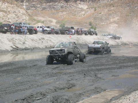 1985 Ford Bronco - Several off-road vehicles navigate a muddy terrain with spectators watching from a sandy embankment.