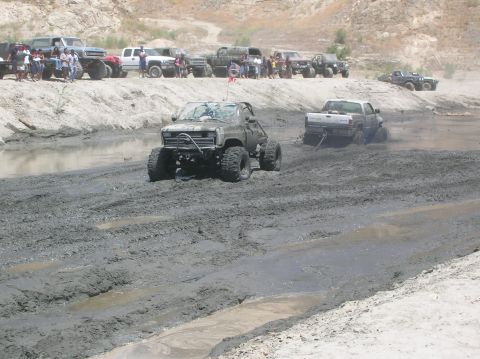 1985 Ford Bronco - Several modified trucks navigate a challenging muddy terrain, with onlookers observing from the side.